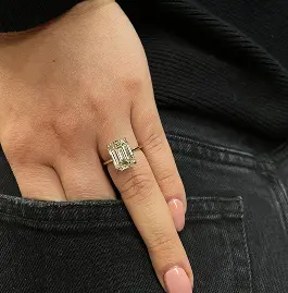 A hand with a gold ring featuring an emerald-cut stone, resting on the edge of a pocket.