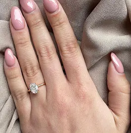 A hand with manicured pink nails wearing a gold ring featuring a large, clear gemstone, resting on a soft, neutral fabric.